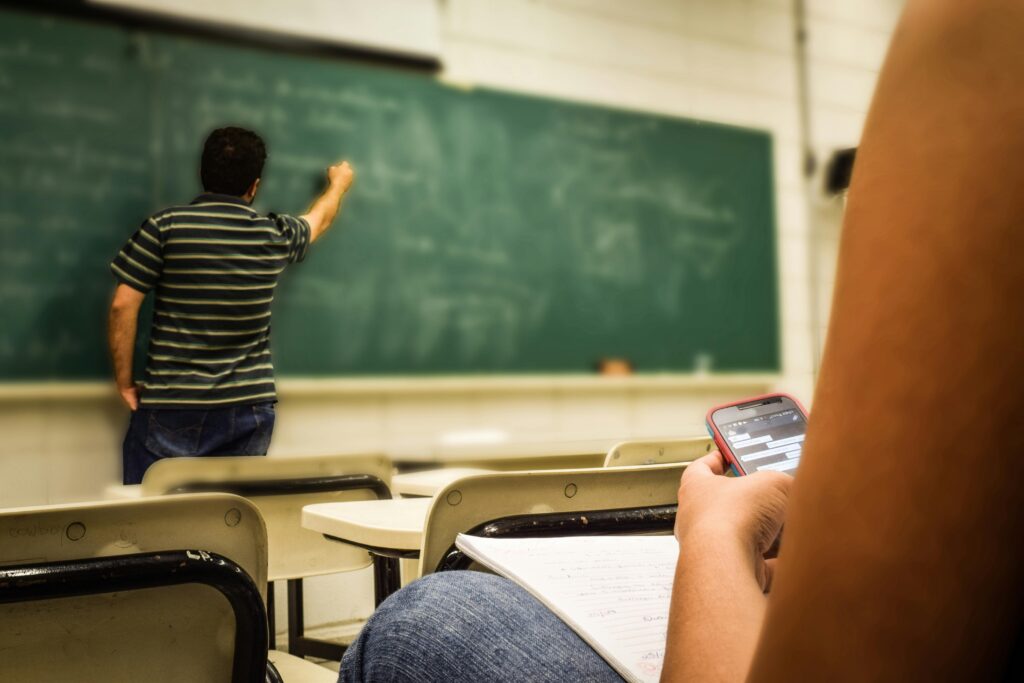 cellular-education-classroom-159844-159844 Student texting in a classroom while teacher is writing on the blackboard.